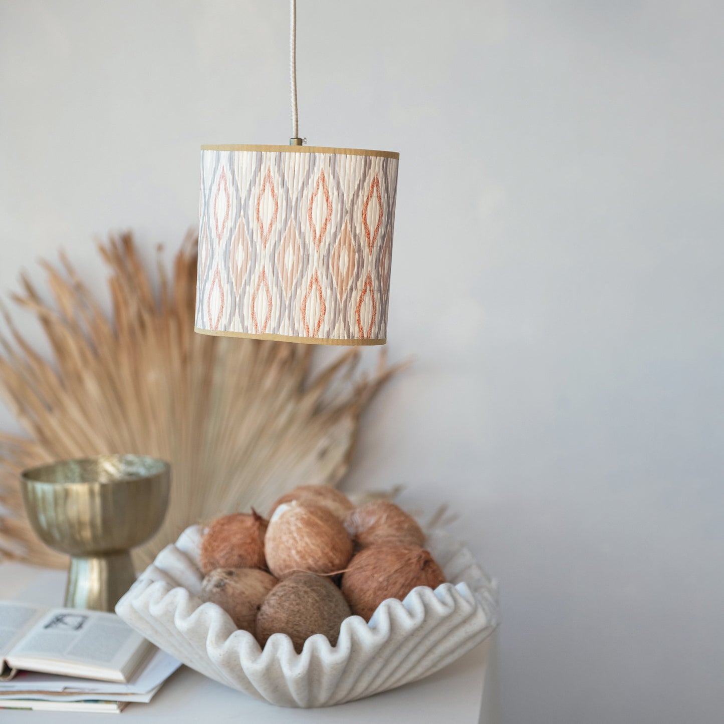 A cylindrical pendant lamp with a beige and orange ikat patterned shade hangs over a styled tabletop scene containing a white fluted bowl of coconuts, a gold cup, and dried pampas grass against a light gray wall.