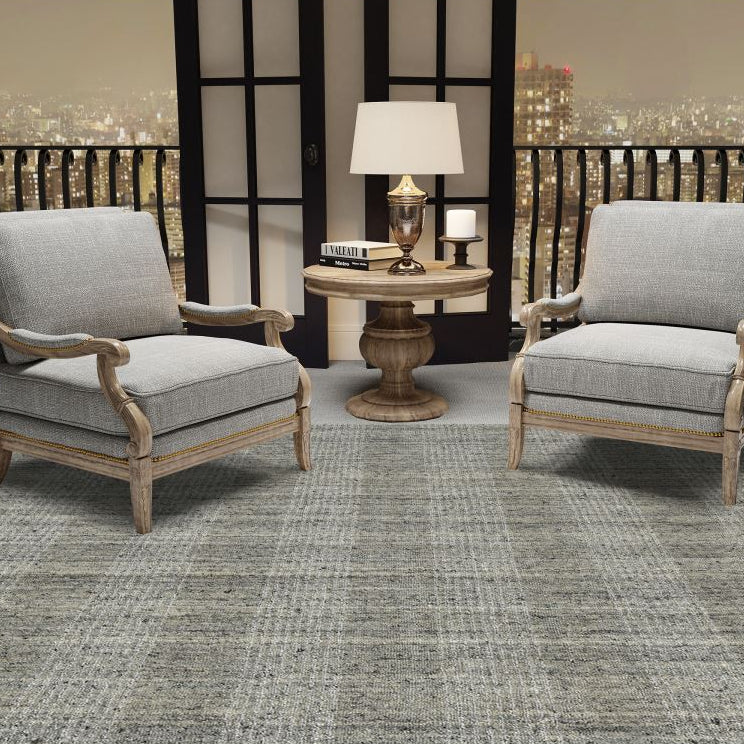 Two gray cushioned chairs with wooden frames on a checkered floor, with a lamp and books on a small round table in the background.