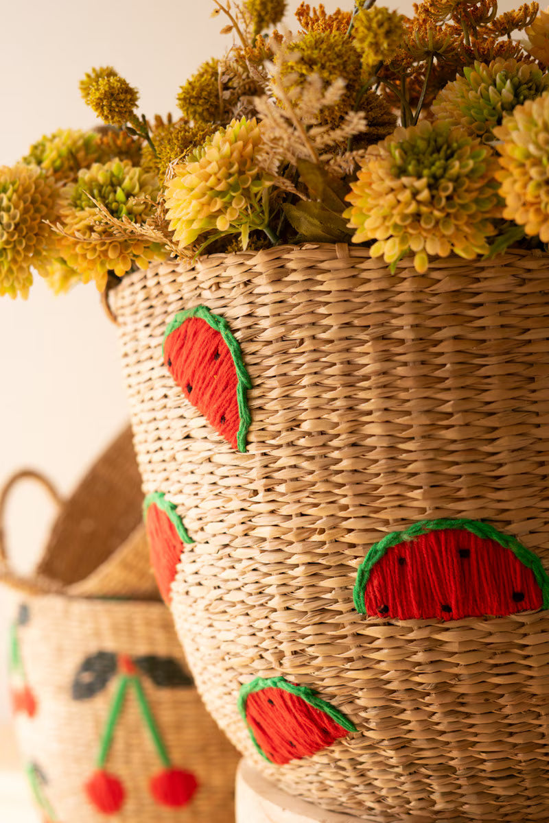 A close-up of a woven straw basket decorated with red and green yarn patterns resembling watermelon slices, filled with dried yellow flowers, with another similar basket blurred in the background.