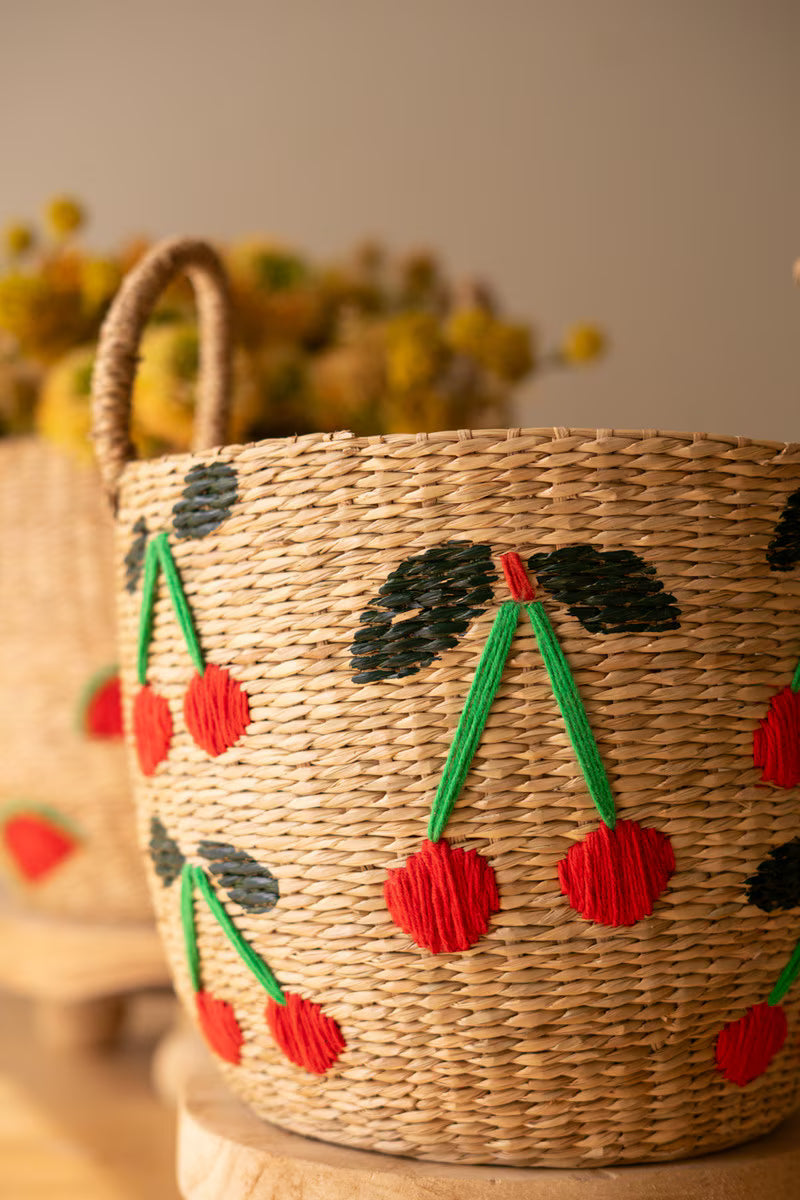 A woven natural fiber basket with a handle, decorated with hand-painted cherry leaves and yarn cherry fruit motifs in red, green, and black, set against a warm neutral background.