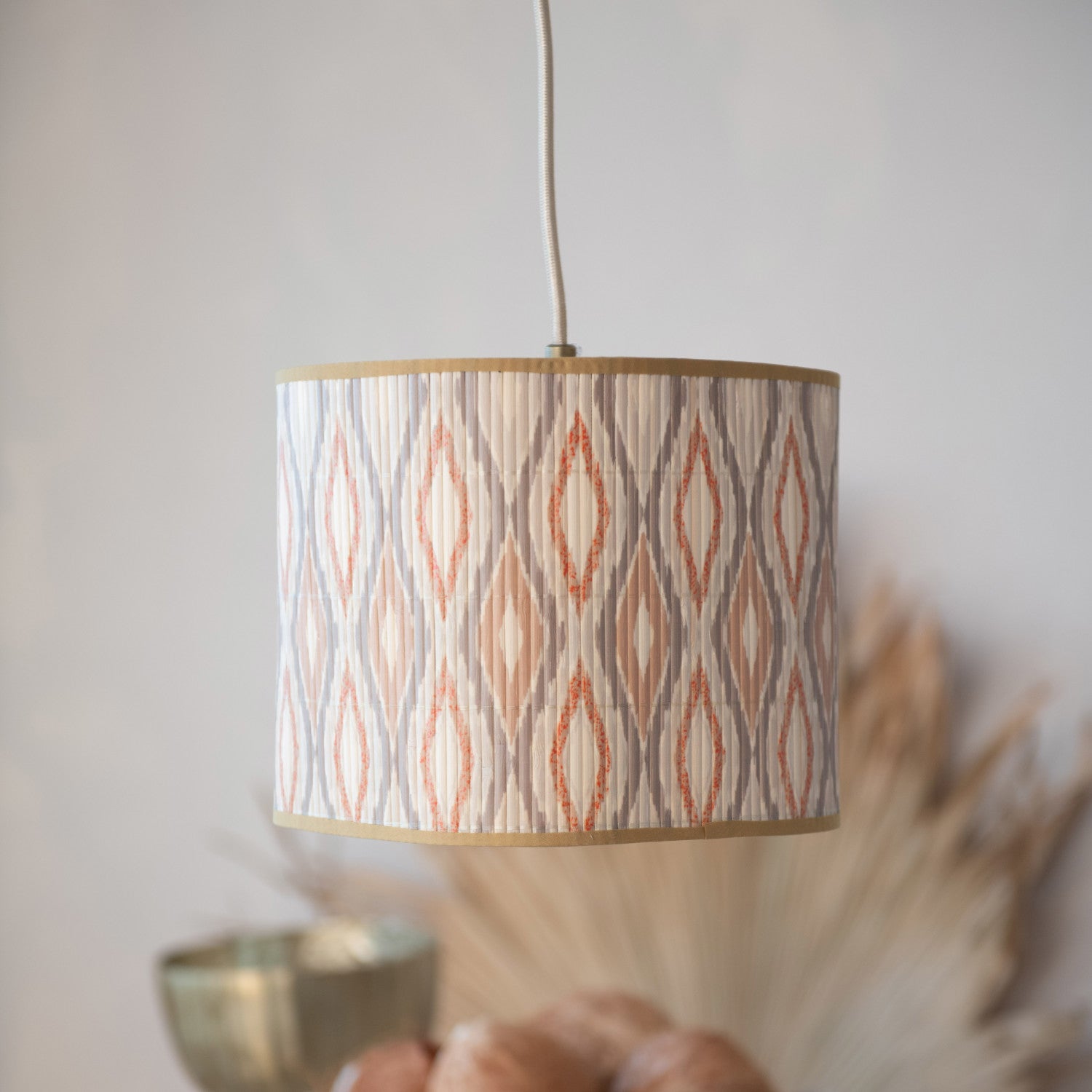 A cylindrical pendant lampshade with a beige, gray, and terracotta ikat pattern, hanging from a white cord against a soft, warm-toned background with blurred decorative elements.