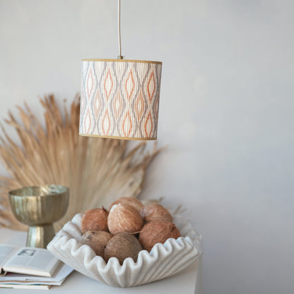 A cylindrical pendant lamp with a beige and orange ikat patterned shade hangs over a styled tabletop scene containing a white fluted bowl of coconuts, a gold cup, and dried pampas grass against a light gray wall.