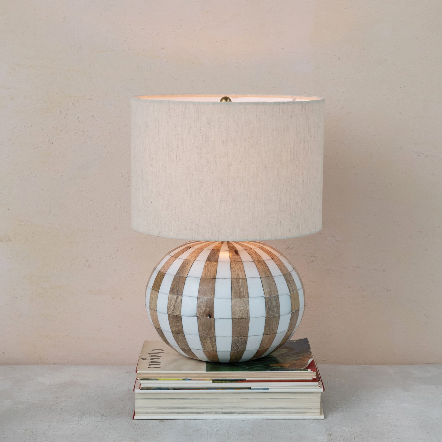 Table lamp with striped base and beige shade on a stack of books against a beige wall.